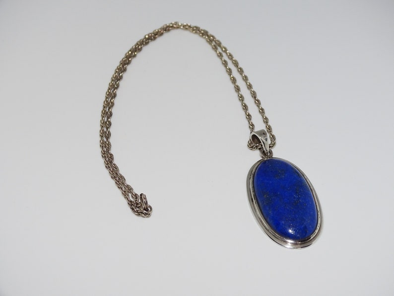 Op de afbeelding: Een zilveren ketting met een grote ovale hanger met een diepblauwe lapis lazuli-steen.