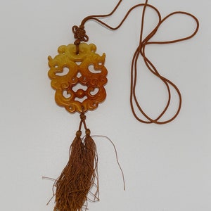Op de afbeelding: Een bruine en gele jade hanger met ingewikkelde gravures hangt aan een bruin koord met kwastjes. De hanger is gevormd als een gezicht met een draaiend ontwerp.