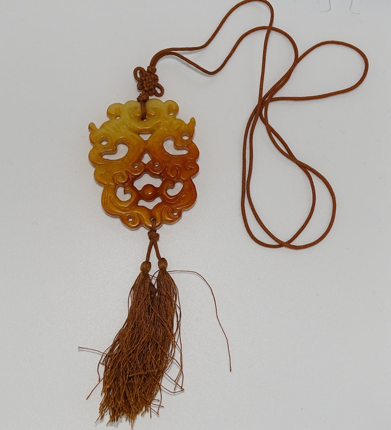 Op de afbeelding: Een bruine en gele jade hanger met ingewikkelde gravures hangt aan een bruin koord met kwastjes. De hanger is gevormd als een gezicht met een draaiend ontwerp.