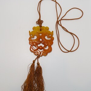 Op de afbeelding: Een gele en oranje jade hanger met ingewikkelde gravures, hangend aan een bruin koord met kwastjes.
