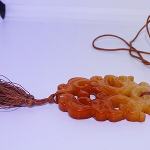 Op de afbeelding: Een gegraveerde jade hanger met een bruine kwast die aan een bruin koord hangt. De hanger is lichtgeel en oranje van kleur en heeft een gedetailleerd ontwerp.
