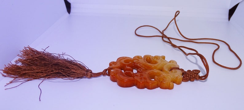 Op de afbeelding: Een gegraveerde jade hanger met een bruine kwast die aan een bruin koord hangt. De hanger is lichtgeel en oranje van kleur en heeft een gedetailleerd ontwerp.