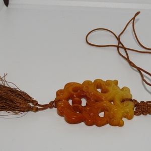 Op de afbeelding: Een gele en oranje jade hanger gegraveerd met een drakenontwerp, hangend aan een bruin koord met kwastjes.