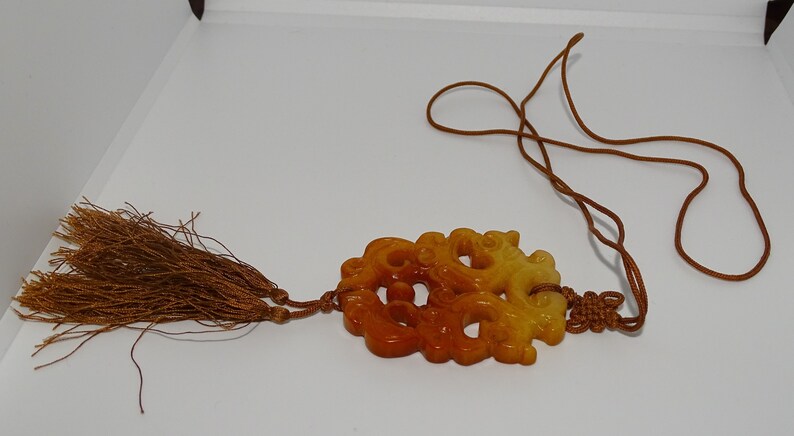 Op de afbeelding: Een gele en oranje jade hanger gegraveerd met een drakenontwerp, hangend aan een bruin koord met kwastjes.