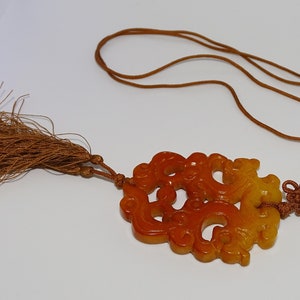 Op de afbeelding: Een gegraveerde amberen hanger met een kwast, met een drakenontwerp. De hanger is bevestigd aan een bruin koord.