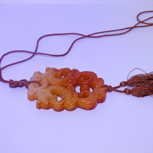 Op de afbeelding: Een bruine en witte jade hanger met ingewikkelde gravures, hangend aan een bruin koord met kwastjes.