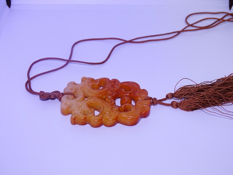 Op de afbeelding: Een bruine en witte jade hanger met ingewikkelde gravures, hangend aan een bruin koord met kwastjes.