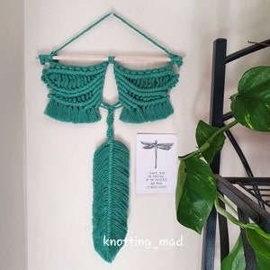 May include: Teal macrame wall hanging with a wooden dowel and a feather design. A card with a dragonfly illustration and text that reads "I ALWAYS TRUST THE DIRECTION OF THE UNIVERSE AND KNOW I'M BEING GUIDED" is also present. The shop name "knotting_mad" is at the bottom.