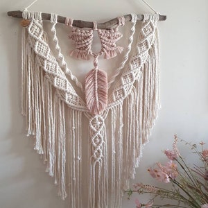 May include: A macrame wall hanging featuring a natural wooden branch and intricate knotting. The design incorporates cream-colored and blush pink cotton cords, with a central feather motif. This handcrafted piece adds a bohemian touch to any space.
