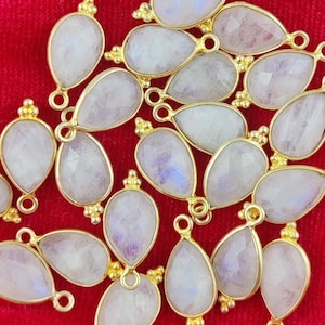 Può includere: Una collezione di 18 ciondoli a forma di lacrima in pietra di luna bianca con anelli di salto dorati. I ciondoli sono disposti in modo casuale su una superficie di velluto rosso.