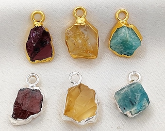 Può includere: Sei ciondoli con pietre preziose nei colori rosso scuro, giallo e turchese. Ogni pietra ha una forma irregolare unica ed è incastonata in una montatura in metallo dorato o argentato con un piccolo anello per appenderla. Presentati su una superficie bianca.