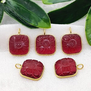 Puede incluir: Cinco colgantes cuadrados con un diseño floral tallado en una piedra preciosa de color rojo intenso. Cada colgante está engastado en un marco dorado y tiene un pequeño lazo para colgar. Los colgantes se muestran sobre una superficie blanca con hojas verdes.