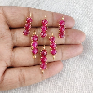 Puede incluir: Varios colgantes dorados, cada uno con una fila vertical de tres o cuatro gemas facetadas de color rosa intenso. Las gemas están espaciadas uniformemente y conectadas por alambre de oro. Los colgantes se muestran sobre un fondo neutro.