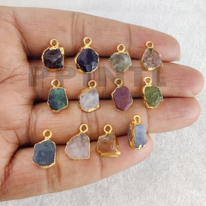 May include: A selection of gemstone pendants in shades of purple, green, and blue. Each pendant is framed with gold-coloured metal and features a small loop for attaching to jewellery. The gemstones have a raw, natural appearance.