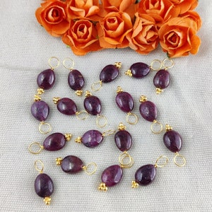 May include: A collection of 18 purple gemstone beads with gold jump rings. Each bead is oval shaped and has a gold jump ring attached to each end.
