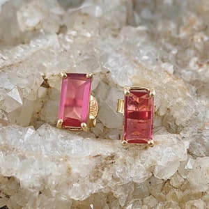 以下が含まれることがあります： ピンクの四角い宝石がついた金のイヤリングのペア。イヤリングは白い背景に置かれています。