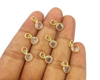 Puede incluir: Varios pequeños colgantes dorados con un centro de piedra preciosa transparente y redondo. Cada colgante tiene un pequeño lazo para sujetarlo a una joya. Los colgantes están sobre una mano, con fondo blanco.