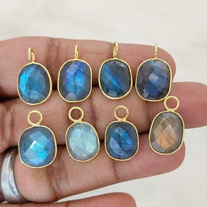 May include: Eight oval-shaped labradorite pendants with faceted surfaces, each set in a gold-toned metal bezel with a small circular jump ring at the top. The stones display iridescent blue and grey tones with a polished finish.