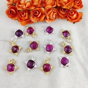 May include: A collection of gemstone pendants with a square, faceted, deep red stone. The pendants have gold and silver-colored metal settings with a starburst design. Orange roses are in the background.