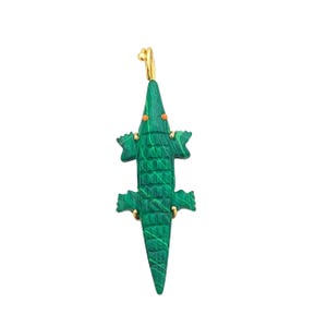May include: A green alligator pendant with gold-colored accents. The pendant is carved with a textured pattern and features orange eyes. The alligator's legs and tail are detailed, and it has a gold-colored loop at the top for hanging.