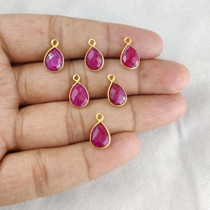 Pode incluir: Seis pingentes em forma de lágrima banhados a ouro com inserções de pedras preciosas vermelhas. Os pingentes estão dispostos em uma mão.