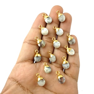 May include: A collection of 14 white and gray agate gemstone pendants with gold-plated bail settings. The pendants are in the shape of a teardrop and are arranged on a hand.
