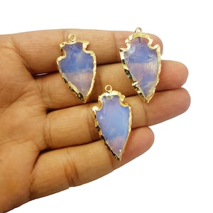May include: Three arrow-shaped gemstone pendants with gold-coloured edging. The gemstones are light blue and white. Each pendant has a small loop at the top for attaching to a chain. The pendants are held in a hand.