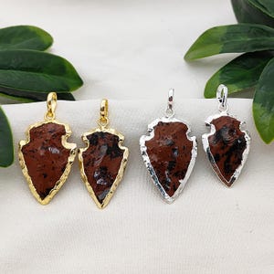 May include: Four arrowhead pendants. Two have gold-colored metal trim and bails, and two have silver-colored metal trim and bails. The arrowheads are a reddish-brown color with black inclusions.