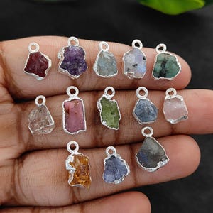 Könnte beinhalten: Eine Sammlung von Anhängern aus rohen Edelsteinen in verschiedenen Farben, darunter Rot, Lila, Grün und Rosa. Jeder Stein ist in eine silberfarbene Metalleinfassung mit einer kleinen Schlaufe zum Aufhängen gefasst. Die Anhänger werden vor einem dunklen Hintergrund gezeigt.