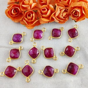 May include: A selection of gold-plated charms featuring faceted, deep magenta gemstones. The charms are square or diamond-shaped, with loops for attaching to jewellery. Orange roses are in the background.