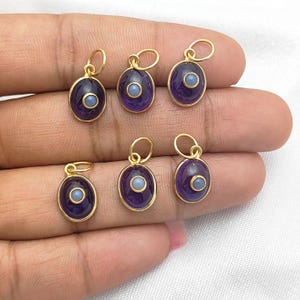 Può includere: Sei ciondoli ovali in oro con una pietra viola e una pietra blu più piccola al centro. I ciondoli sono su una mano.