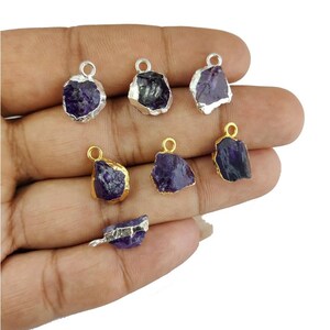 May include: A collection of amethyst gemstone pendants with silver and gold-colored metal accents. The raw, irregular amethyst stones are a deep purple color. Each pendant has a small loop for attaching to a chain or other jewelry component.
