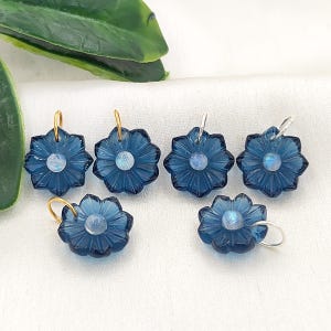 May include: Six flower-shaped pendants carved from deep blue translucent material, each featuring a small iridescent round stone at the center. Two pendants have gold-toned metal jump rings, while four have silver-toned metal jump rings attached.