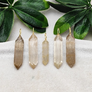 May include: Five crystal pendants with gold-colored loops. The crystals vary in size and shade, ranging from clear to light brown. The pendants are displayed on a white surface with green leaves in the background. These are ideal for jewelry making.