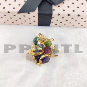 May include: A collection of raw gemstone pendants in various colors, including green, purple, and gray, with gold-colored metal edging. The pendants are displayed on a white surface, with a pink gift box with black polka dots and a black ribbon in the background.