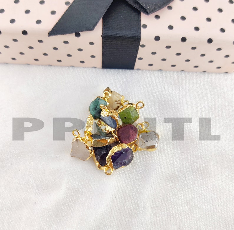 May include: A collection of raw gemstone pendants in various colors, including green, purple, and gray, with gold-colored metal edging. The pendants are displayed on a white surface, with a pink gift box with black polka dots and a black ribbon in the background.