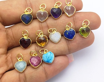 May include: A collection of heart-shaped gemstone pendants in various colors, including blue, purple, and green, with gold-colored metal settings. Each pendant has a small loop at the top for attaching to a chain or bracelet. The gemstones are faceted, giving them a sparkling appearance.