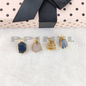 May include: Four gemstone pendants with gold-colored metal edging. The gemstones are various colors, including blue, tan, and pink. Each pendant has a small loop at the top for attaching to a chain. The pendants are displayed on a white surface.