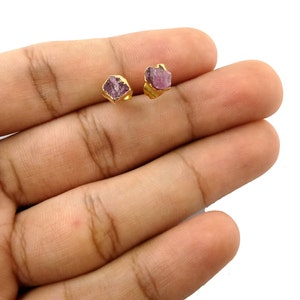 Puede incluir: Un par de pendientes de botón dorados, cada uno con una piedra preciosa de color púrpura claro y corte rugoso. Los pendientes están engastados en metal dorado, sobre un fondo blanco.