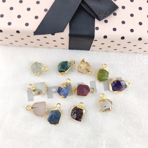 May include: A collection of raw gemstone pendants with gold-colored metal edging. The gemstones are various colors, including green, blue, purple, and clear. Each pendant has a small loop for attaching to a chain or cord. The background includes a pink gift box with black polka dots.