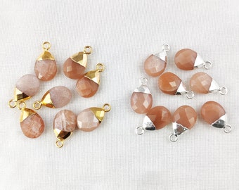 May include: A collection of 12 teardrop-shaped sunstone gemstone pendants with gold and silver plated jump rings. The stones are a light peach color with a shimmery finish.