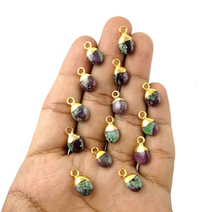 May include: A collection of 14 green and purple gemstone pendants with gold-plated tops. The pendants are shaped like teardrops and are arranged on a hand.