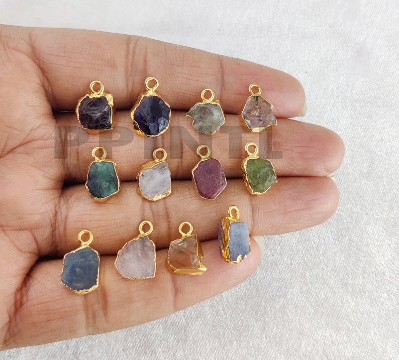 May include: A collection of gemstone pendants in various colors, including purple, green, and blue. Each pendant has a gold-colored metal border and a small loop for attaching to a chain or other jewelry. The gemstones have a rough, natural appearance.