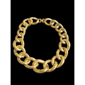 May include: A gold-toned chunky chain necklace featuring large, interlocking oval links with a polished, reflective surface. The necklace is secured with a rectangular clasp mechanism at the top, creating a continuous circular shape.
