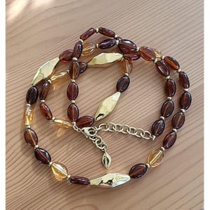 May include: A necklace featuring oval-shaped beads in varying shades of translucent amber and dark brown, interspersed with polished gold-tone metal diamond-shaped accents and small gold-tone spacer beads, finished with a gold-tone lobster clasp.