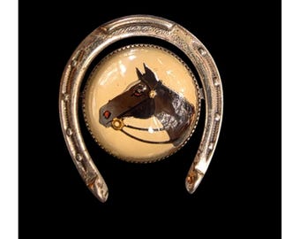 May include: A vintage horse-themed brooch. The brooch features a detailed painting of a horse's head within a circular frame, set inside a silver horseshoe. The horse is brown with a black mane and a red accent on its nose. The background is a light cream color.