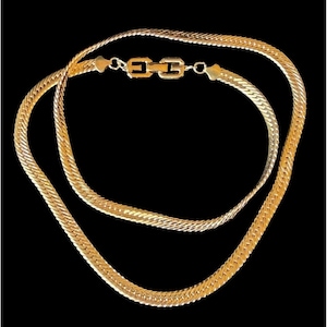 Puede incluir: Un collar dorado con un diseño de cadena de espiga. El collar presenta un cierre rectangular con un diseño decorativo. La joya se muestra sobre un fondo negro.