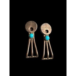 May include: A pair of silver-toned earrings with a Native American design. Each earring features a circular top with a geometric pattern, a turquoise stone, and a triangular base with three vertical bars. The earrings are set against a black background.