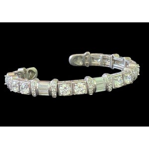 May include: A silver-toned cuff bracelet featuring a repeating pattern of round-cut and baguette-cut clear stones set in metal links. The bracelet has a hinged opening mechanism with heart-shaped details at the ends of the band.