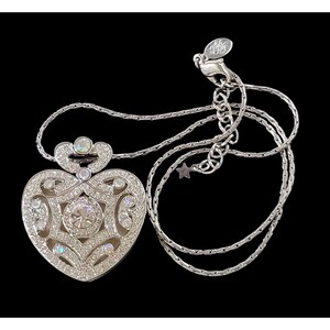 May include: Silver-toned heart pendant necklace with clear crystals. The heart-shaped pendant features a central large crystal surrounded by smaller crystals and ornate detailing. The necklace has a delicate chain and a lobster clasp.